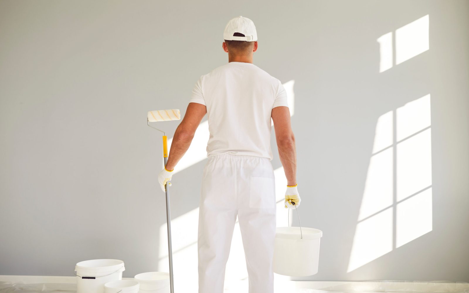 Back view of a male painter in white uniform with bucket and paint roller in hands going painting a grey wall in empty room. Renovation, apartment repair and renovating house concept.